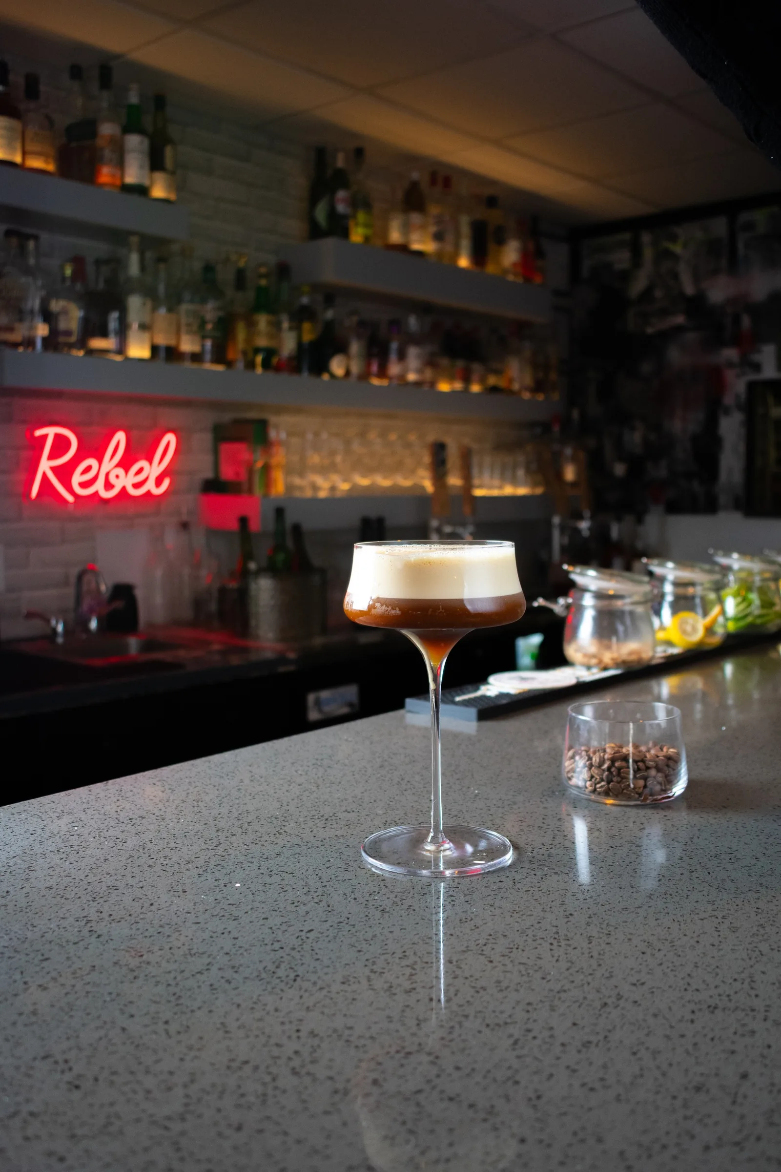 A coupe of espresso martini on the bar, the red Rebel neon burning behind it
