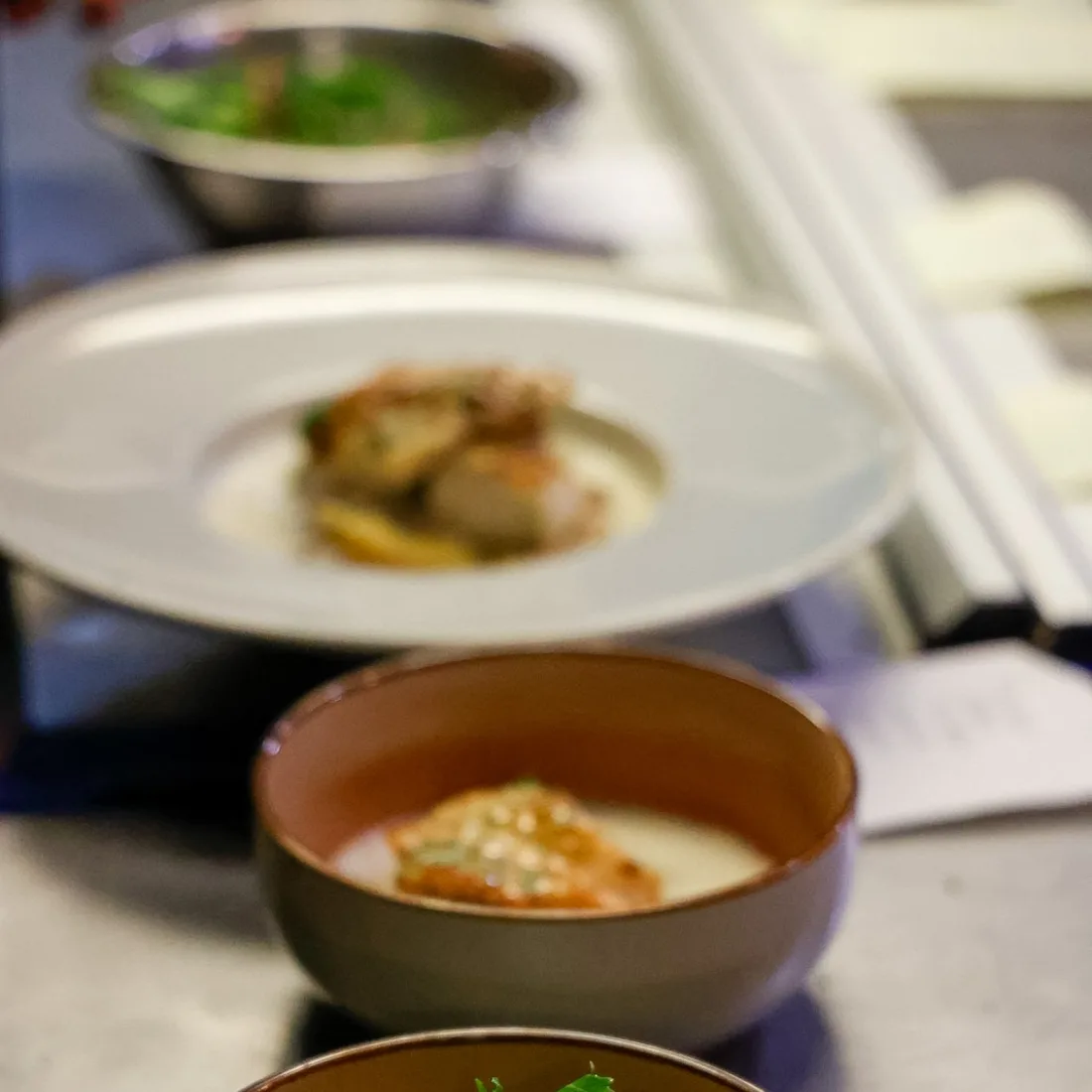 Three bowls of a seafood course lined up on the stainless pass, ready to go out
