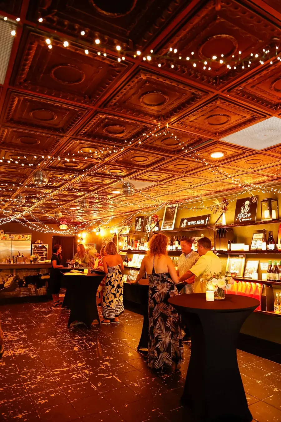 Guests standing at cocktail tables by the wine wall, string lights across the pressed-tin ceiling above