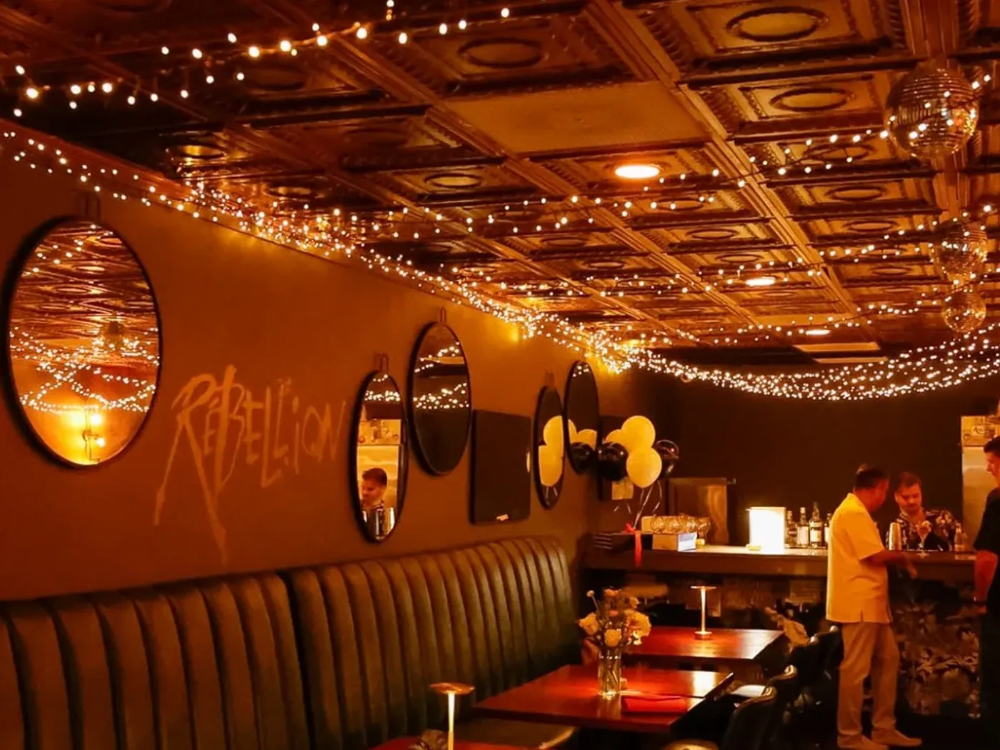The private room laid for dinner — green banquette, candlelit tables, the Rebellion mark on the wall under a pressed-tin ceiling